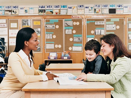 Teacher Sitting at a School Desk Showing a Book to a Parent and Her Son